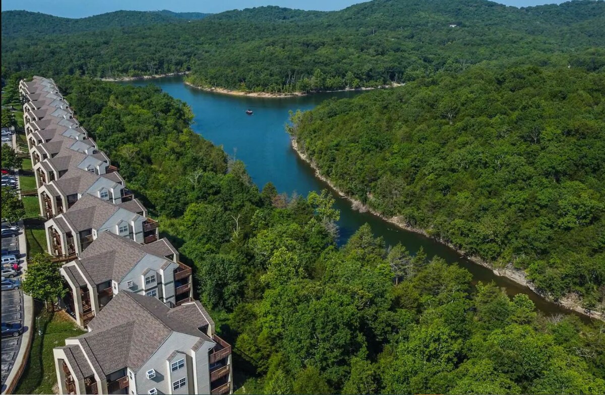 An aerial view captures the lush greenery surrounding the tranquil waterway, contrasting with the architectural lines of the nearby condos. The natural landscape blends seamlessly with the buildings, emphasizing the serene setting of the resort nestled in the Ozark Mountains.