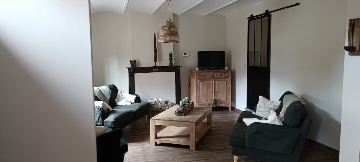 A living area features a wooden coffee table surrounded by comfortable seating. A natural wood cabinet and a television are positioned against the wall. Textured light-colored walls and a ceiling with soft curves enhance the spacious feel of the room.