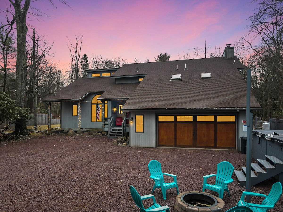 The exterior of the cottage is captured during twilight, showcasing a spacious layout with a sloping roof. The garage doors and large windows provide a warm glow, while brightly colored chairs surround a fire pit in the foreground, inviting relaxation amid the natural surroundings.