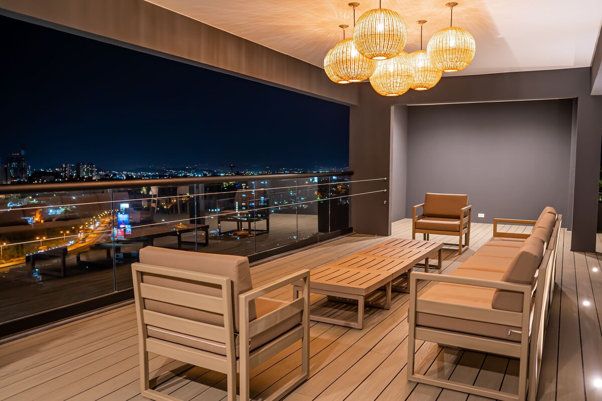 An outdoor lounge area features modern furniture with a wooden coffee table and comfortable seating. Large windows provide expansive views of the city at night. Three decorative light fixtures hang from the ceiling, casting a warm glow over the space.
