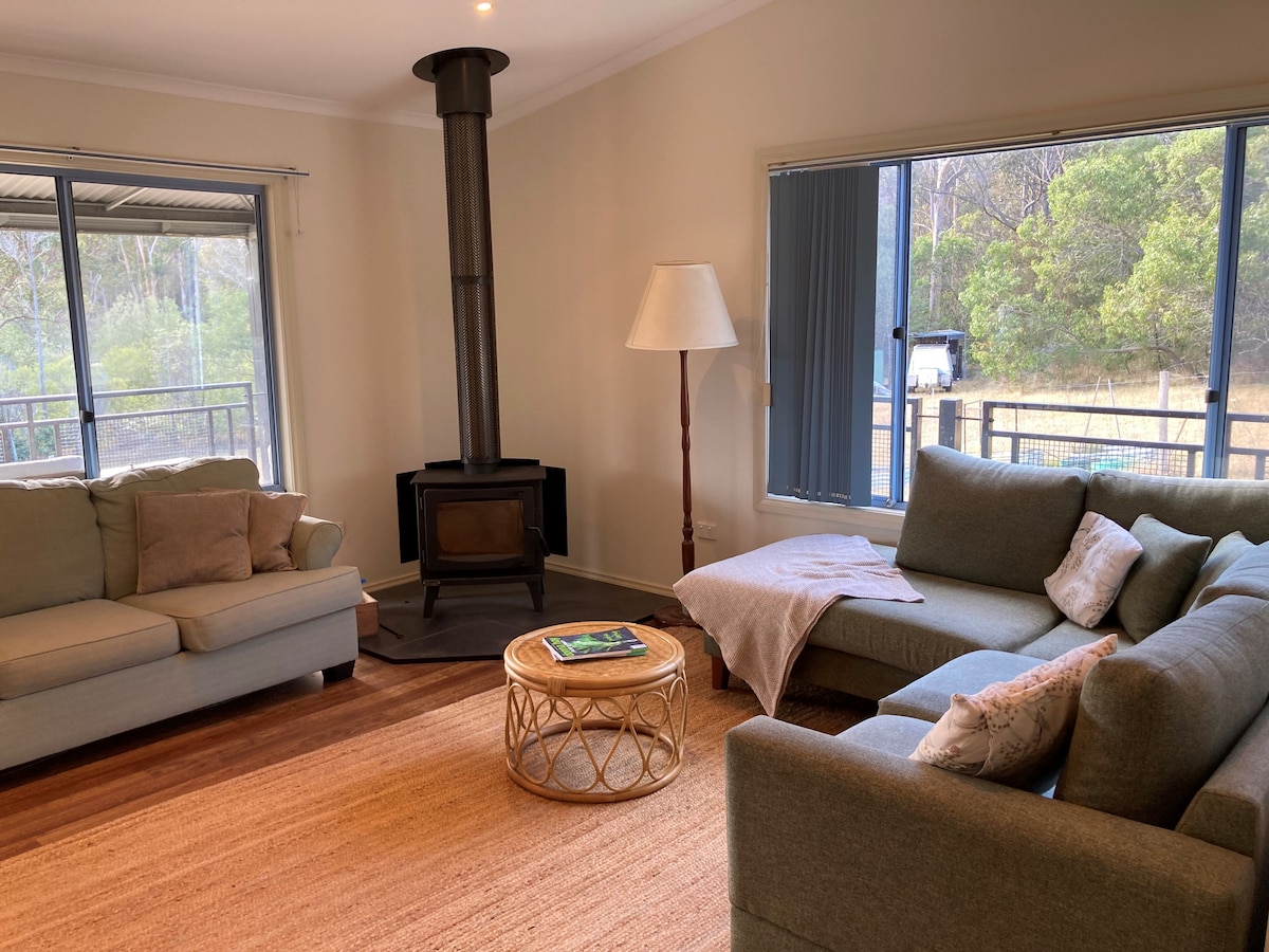 The living area features a warm ambiance with comfortable seating on two sofas and a round coffee table. A wood-burning stove stands against a wall, while large windows allow natural light to fill the space and provide views of the surrounding greenery.