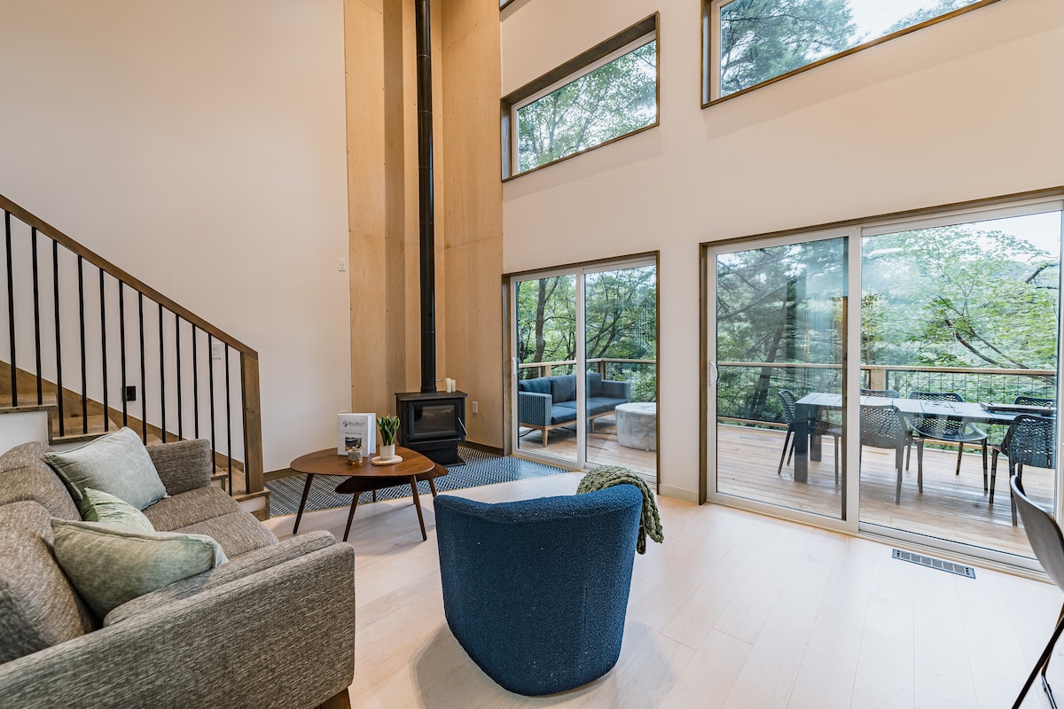 A spacious living area features floor-to-ceiling windows, allowing natural light to fill the space. A comfortable sofa and accent chair are positioned around a small table. A wood-burning stove is visible, and an outdoor deck with dining chairs can be seen through the glass doors.