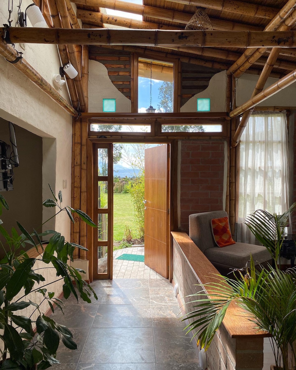 The interior features a welcoming entrance with wooden doors opening to a green landscape outside. Natural light floods the space, highlighting a blend of rustic materials and modern design. Potted plants add a touch of greenery, enhancing the overall serene atmosphere.