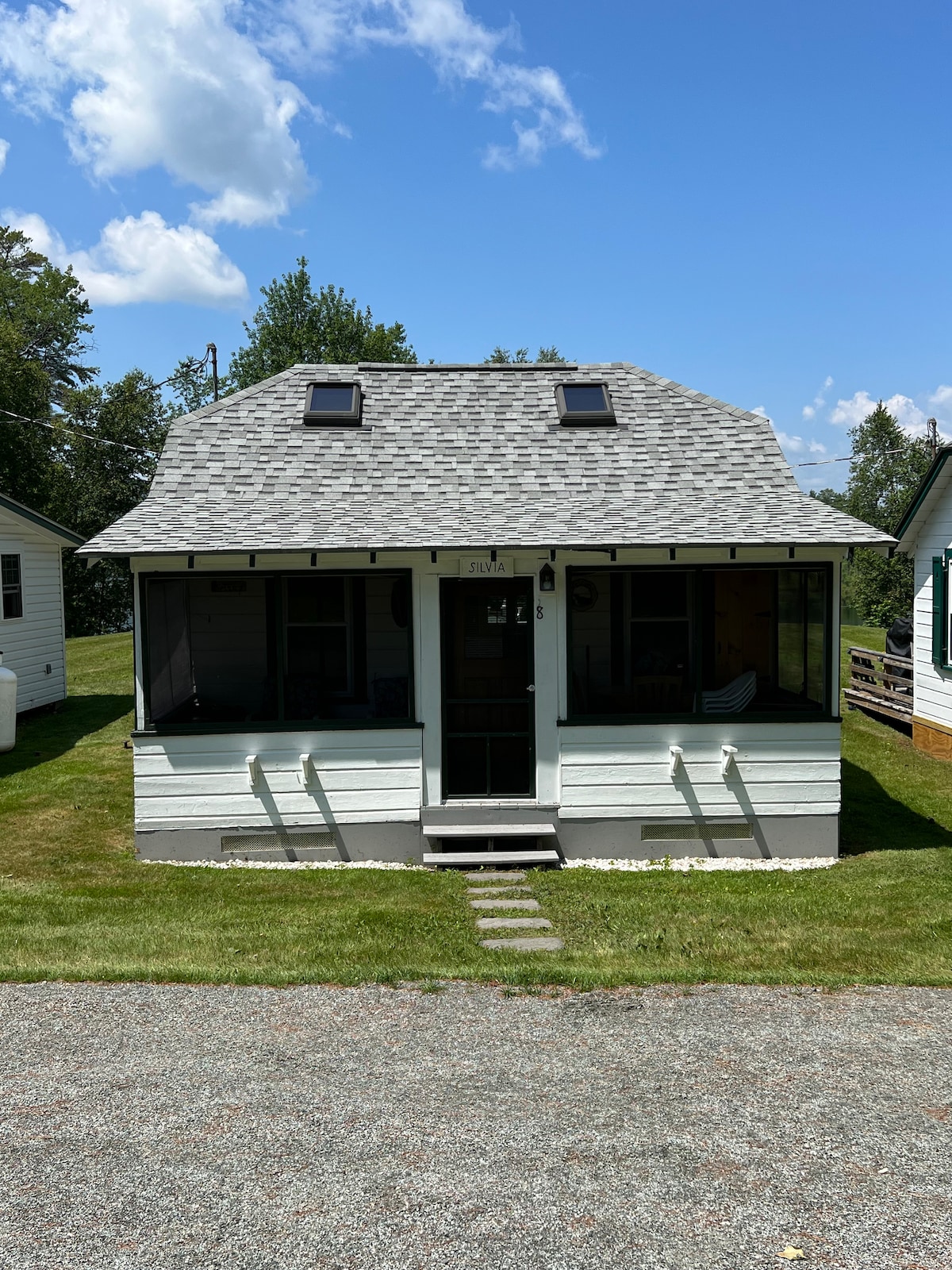 The cottage features a charming exterior with a gabled roof and dual skylights. A welcoming screened-in porch is visible, providing ample seating and a table for dining. The grassy yard complements the inviting facade of the property, enhancing the overall serene atmosphere.