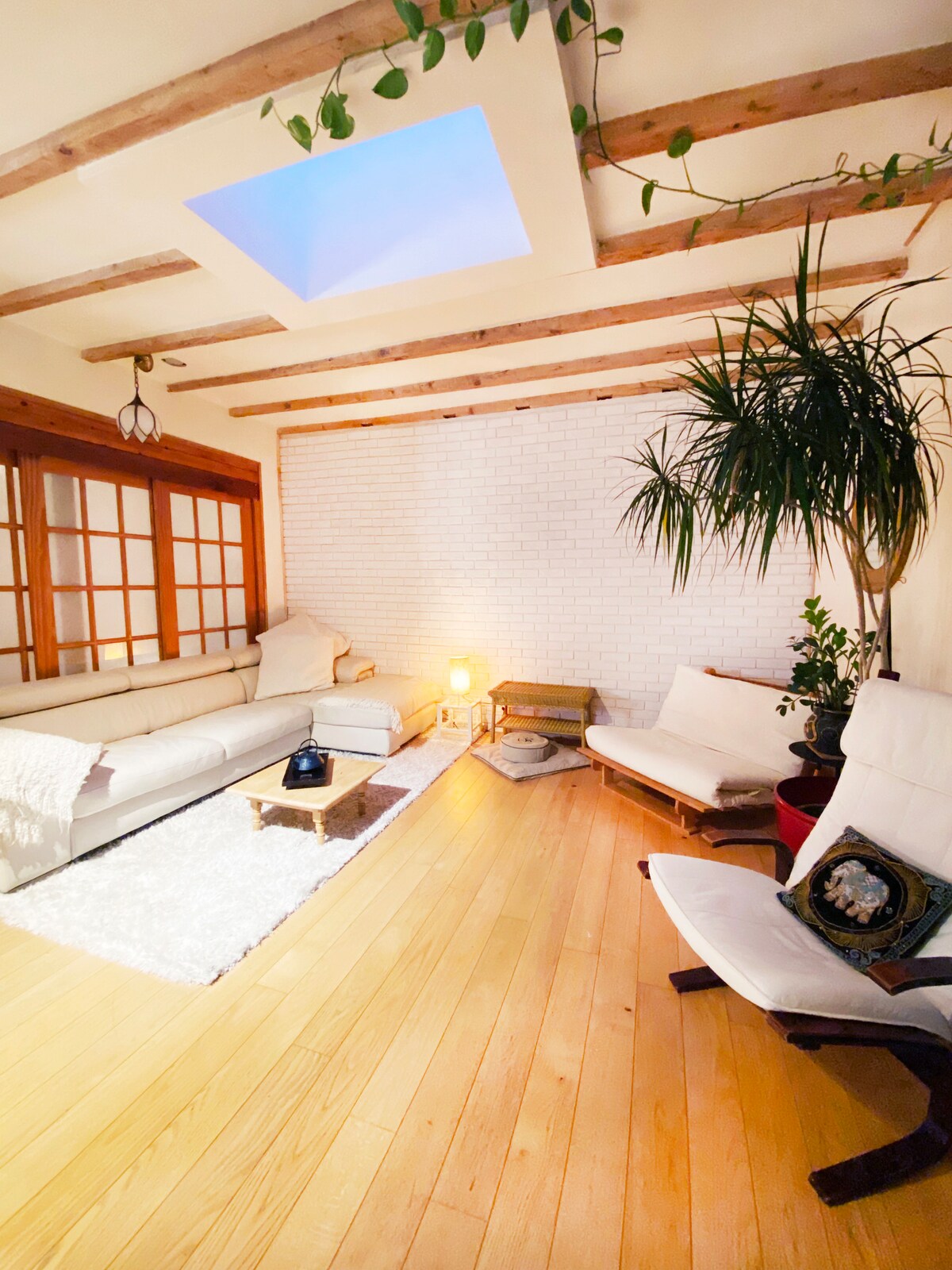 A cozy living area is highlighted by natural wood beams and a skylight, allowing soft light to filter in. White seating is arranged around a low table on a light rug. Potted plants add a touch of greenery, enhancing the space's relaxing atmosphere.