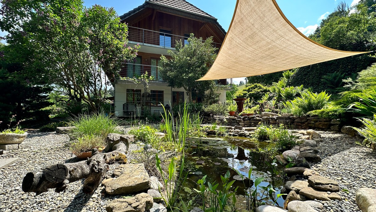 A serene garden surrounds the house, featuring a peaceful pond adorned with water plants and smooth stones. Natural shade is provided by a fabric canopy overhead, while lush greenery and gravel pathways add to the tranquil atmosphere. The home’s structure stands in the background.