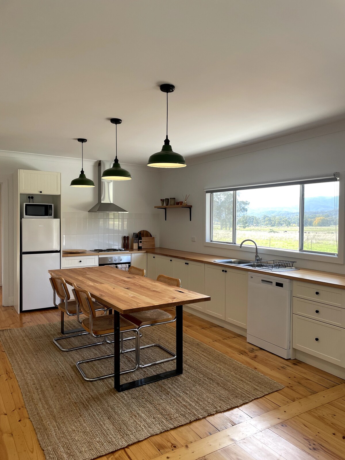 The kitchen features modern appliances and a wooden dining table surrounded by metal-framed chairs. Green pendant lights hang above the table, illuminating the space. A large window offers views of the surrounding mountains, while natural light enhances the neutral decor.