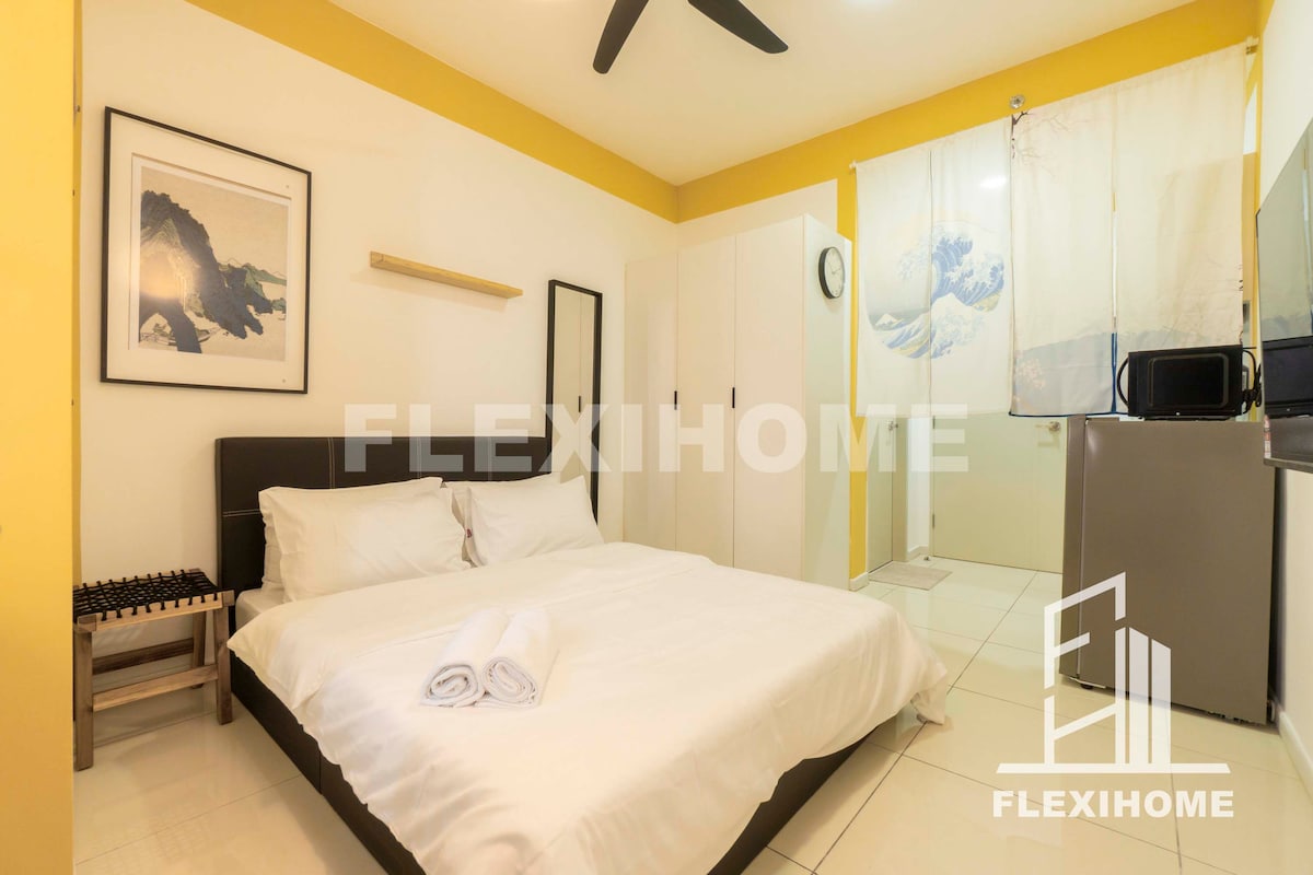 The spacious sleeping area features a comfortable bed with neatly arranged linens and towels. A minimalist design is evident with a wall-mounted shelf and art piece. A closet and kitchenette are visible in the background, while a decorative screen adds privacy.