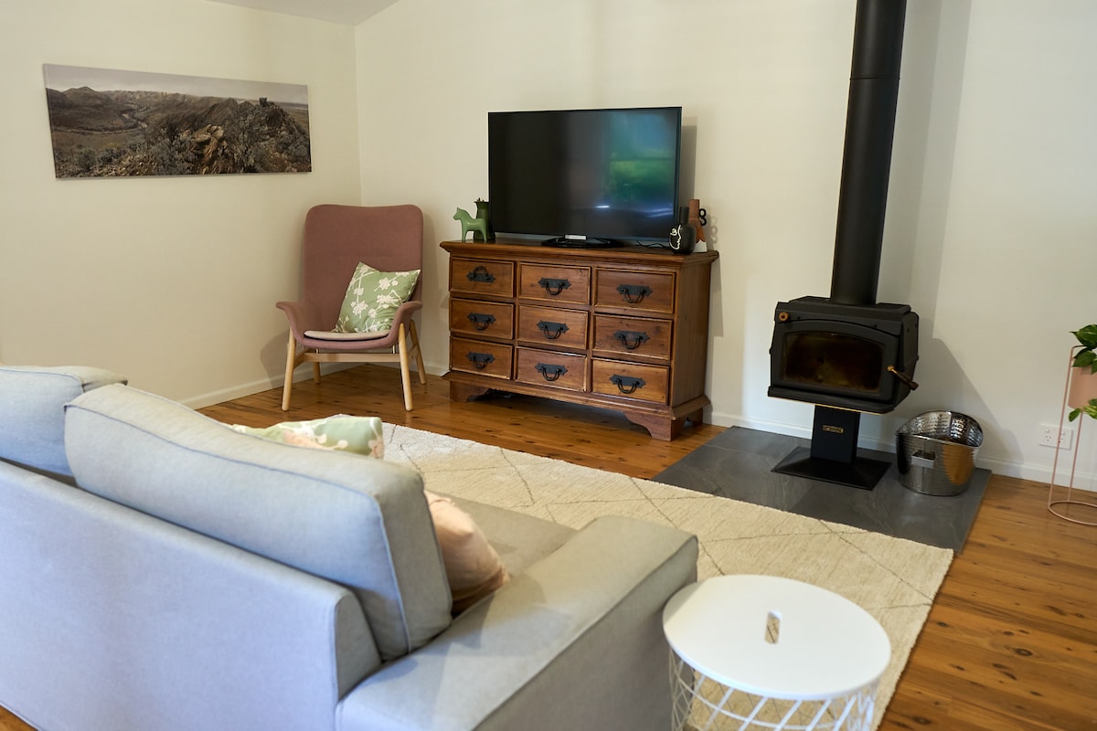 A serene living area features a soft gray sofa positioned opposite a wooden cabinet with a flat-screen TV. A rustic wood stove stands nearby, accompanied by a stylish accent chair with a pastel cushion. A textured rug adds warmth to the hardwood floor.