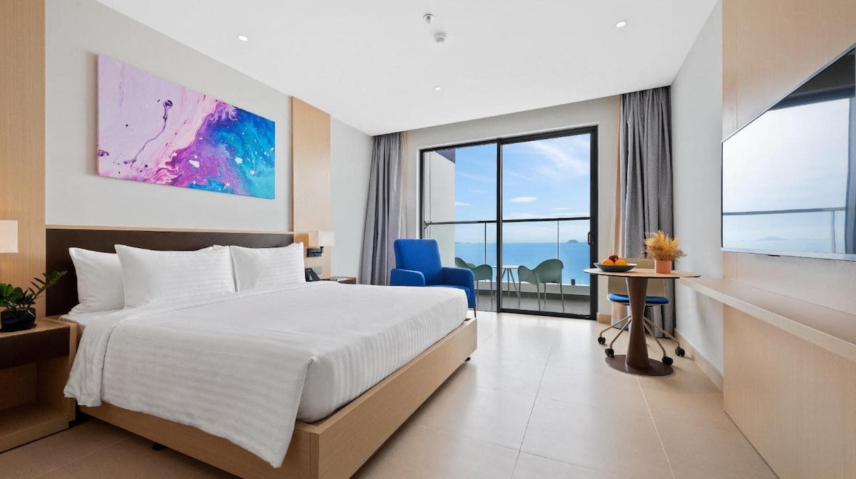 A modern bedroom features a spacious bed with crisp white linens, complemented by an abstract artwork above. A seating area includes a blue chair and a small table with fruit. Large glass doors offer a view of the ocean and access to a private balcony.