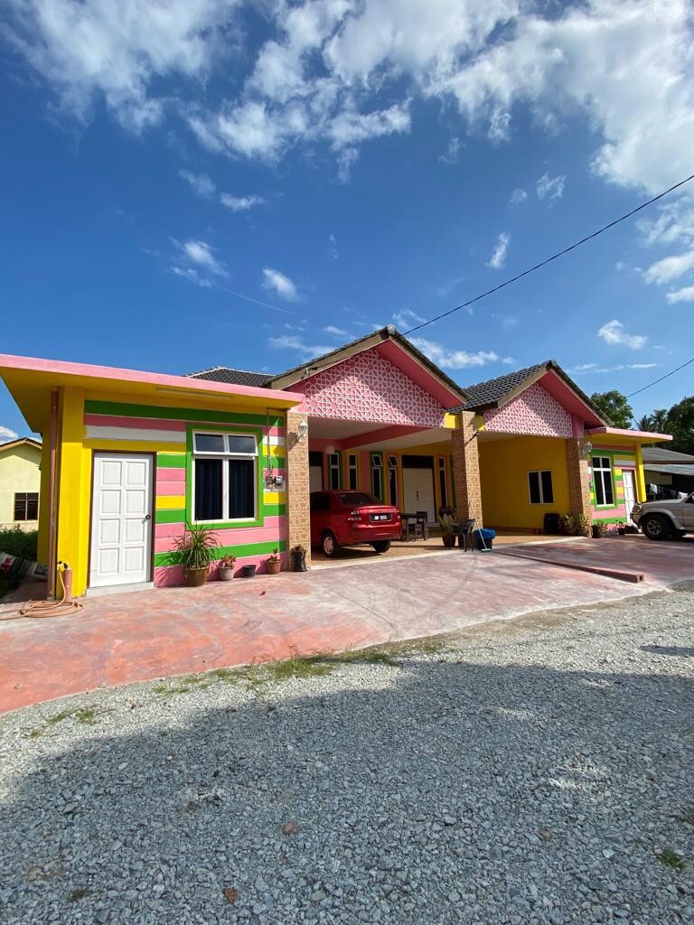 A colorful building exterior features bright yellow and pink walls, with decorative stripes adding visual interest. The driveway is paved with light-colored stones, and several vehicles are parked in front. A clear blue sky with scattered clouds complements the cheerful setting.