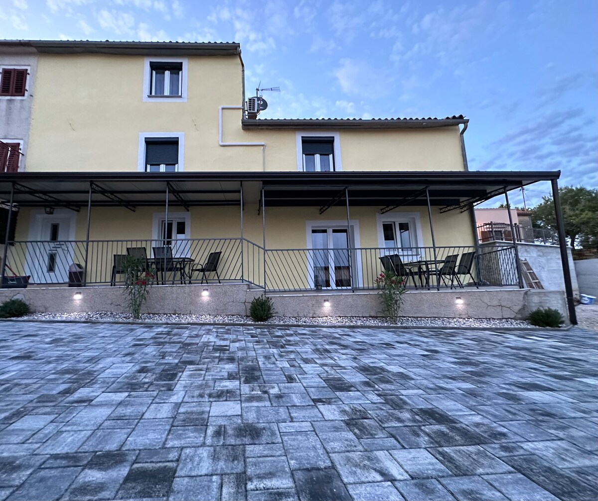 The exterior of the house features a covered terrace with multiple tables and chairs arranged for outdoor seating. A spacious stone-paved area surrounds the property, while white walls and dark shutters provide a harmonious contrast to the light blue sky above.