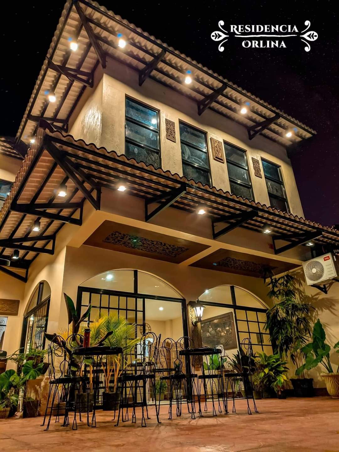 An elegant exterior view of the building showcases multiple large windows adorned with decorative patterns. Soft lighting illuminates the structure, highlighting its architectural features. A courtyard area is visible, furnished with several iron chairs surrounded by lush greenery, creating a welcoming outdoor space.