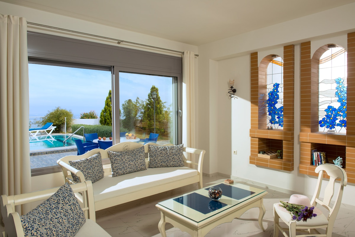 A serene living area features a light-colored sofa adorned with patterned cushions, complemented by a glass coffee table. Large windows offer a view of an outdoor pool area, framed by lush greenery. Decorative elements include stained glass windows with floral designs.
