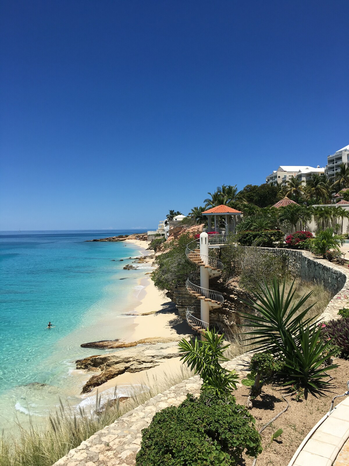 A coastline is depicted with soft sand and clear turquoise waters gently lapping the shore. Lush greenery and colorful flowers frame the scene, while a staircase winds toward a viewing platform overlooking the beach. The sky is bright blue, complementing the serene landscape.