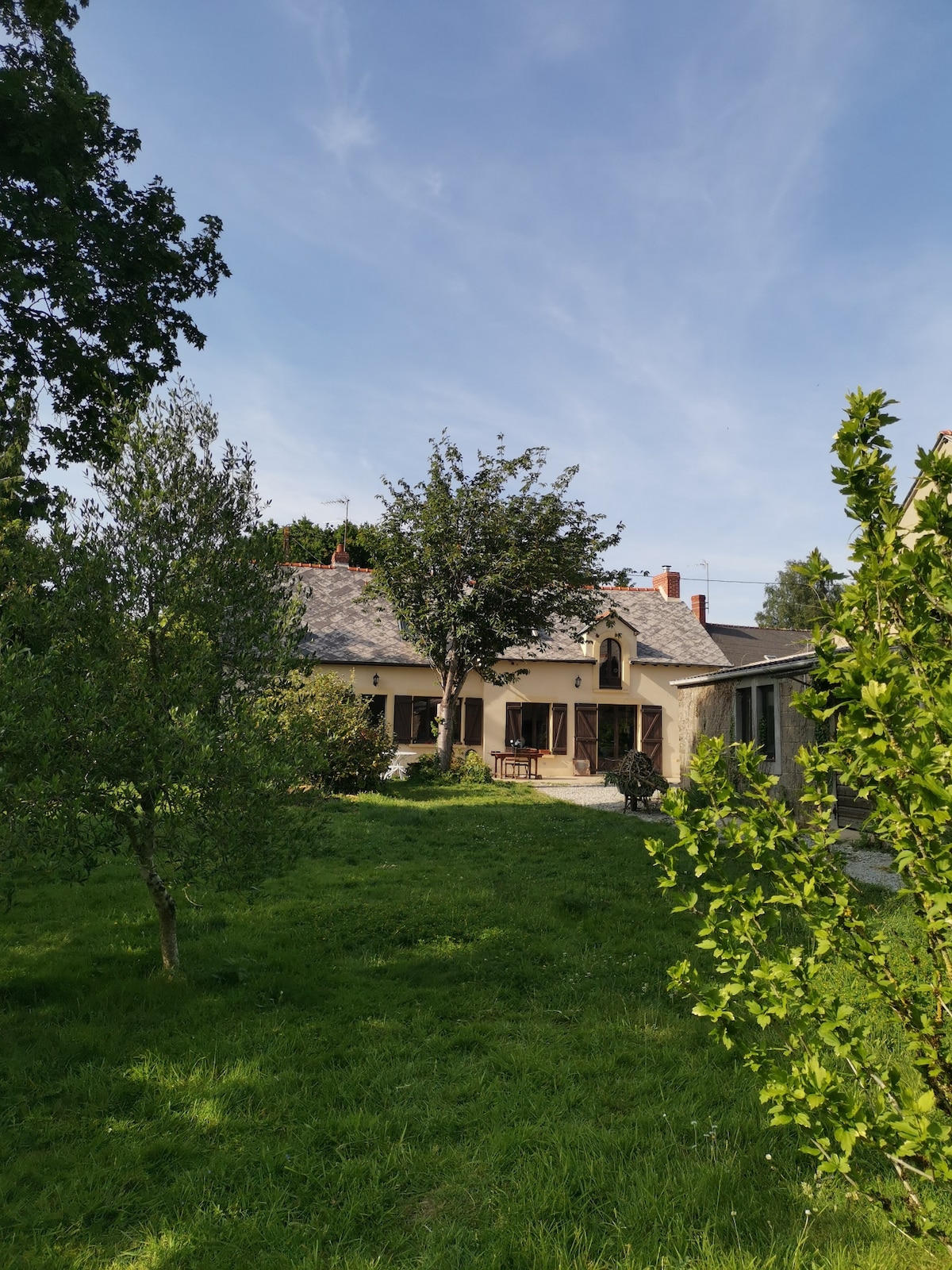 The exterior of a renovated longère is visible, framed by lush greenery. A spacious garden surrounds the house, featuring trees and a well-maintained lawn. The home, with a light-colored facade, shows large windows that invite natural light. A tranquil setting is emphasized by the clear blue sky.