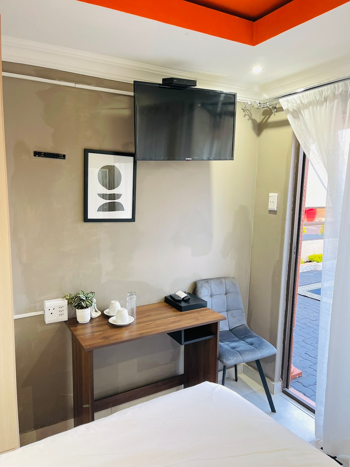 A compact sitting area is visible, featuring a small table and a gray chair. A flat-screen television is mounted on the wall, alongside a framed artwork. Natural light enters through a large window, which is draped with sheer white curtains.