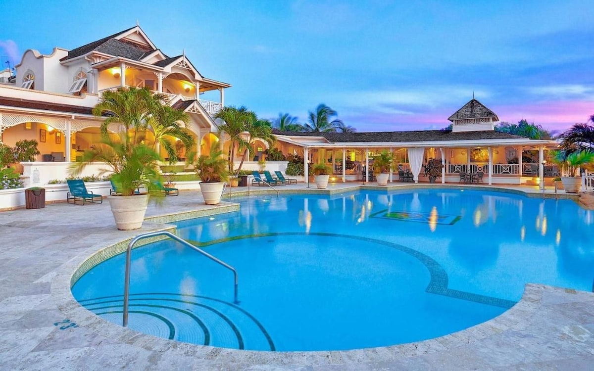 A large infinity pool is viewed with inviting turquoise waters, surrounded by palm trees and lounge chairs. The two-story penthouse structure features a wide patio area with seating. The evening sky is filled with vibrant pink and purple hues, enhancing the serene atmosphere of the resort.