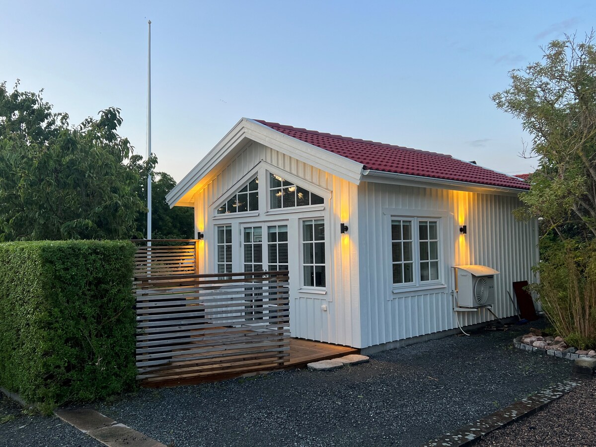 A charming white cottage with a red roof is positioned amidst greenery. Large windows allow natural light to fill the interior. Soft lighting enhances the exterior features, while a gated wooden fence provides a sense of privacy. Gravel pathways lead up to the entrance.