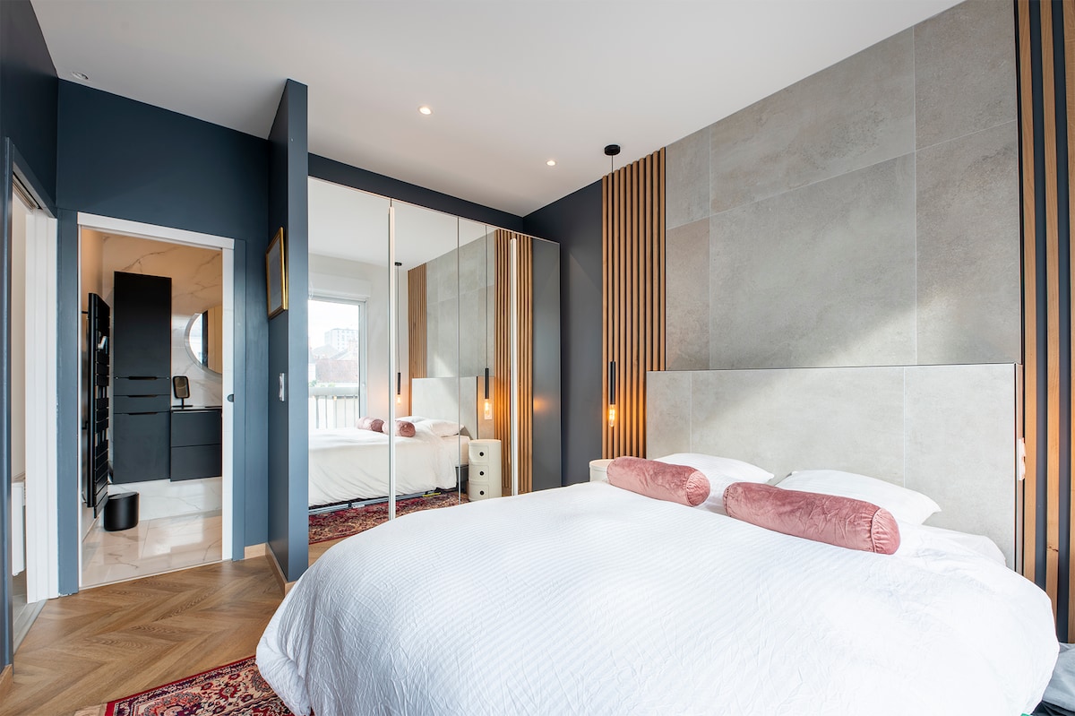 A modern bedroom features a spacious bed adorned with soft pillows. Natural light is allowed to enter through large windows. A stylish mirrored wardrobe is positioned alongside a textured wall. An open doorway leads to a well-lit bathroom area, contributing to a cozy and balanced atmosphere.