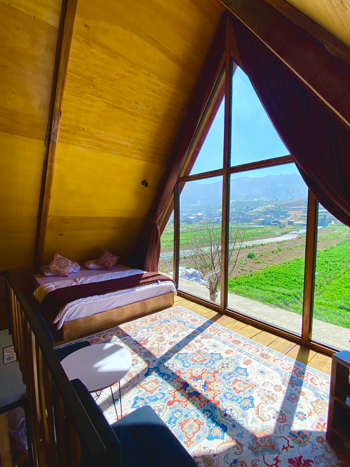 A serene loft space is highlighted by a large triangular window, providing sweeping views of lush greenery and distant mountains. A cozy bed is positioned against the wall, with patterned pillows adding a touch of comfort. A round table sits beside the bed on a soft area rug.
