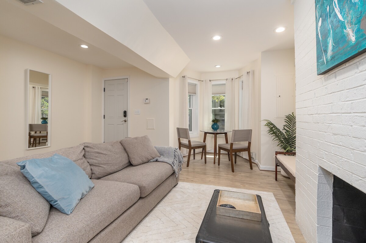 A comfortable living space features a soft gray couch with decorative pillows. A small dining table with two chairs is positioned by a window, allowing natural light to illuminate the room. The corner fireplace adds a cozy element to the overall design.