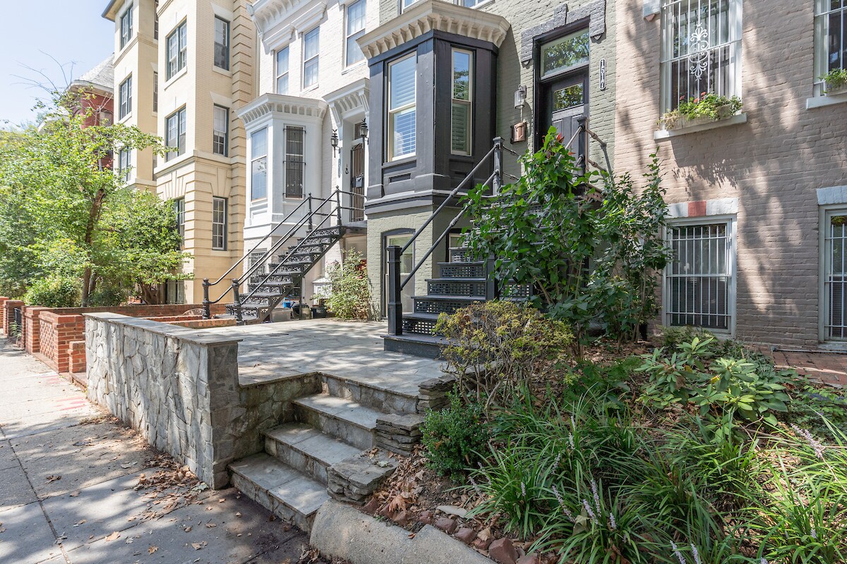 A charming townhouse exterior is displayed, featuring a stone walkway leading to a separate entrance. Lush greenery and plants surround the entryway, and the townhouse showcases distinct architectural elements, including steps and a decorative railing.