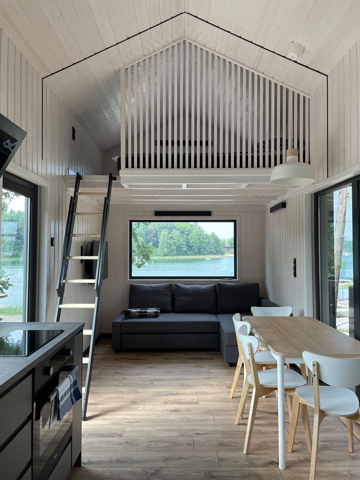The interior of a tiny house showcases a ladder leading to a lofted sleeping area. A dark sectional couch is positioned next to a dining table with white chairs. Large windows allow natural light and views of the lake, while light wooden flooring enhances the spacious feel.