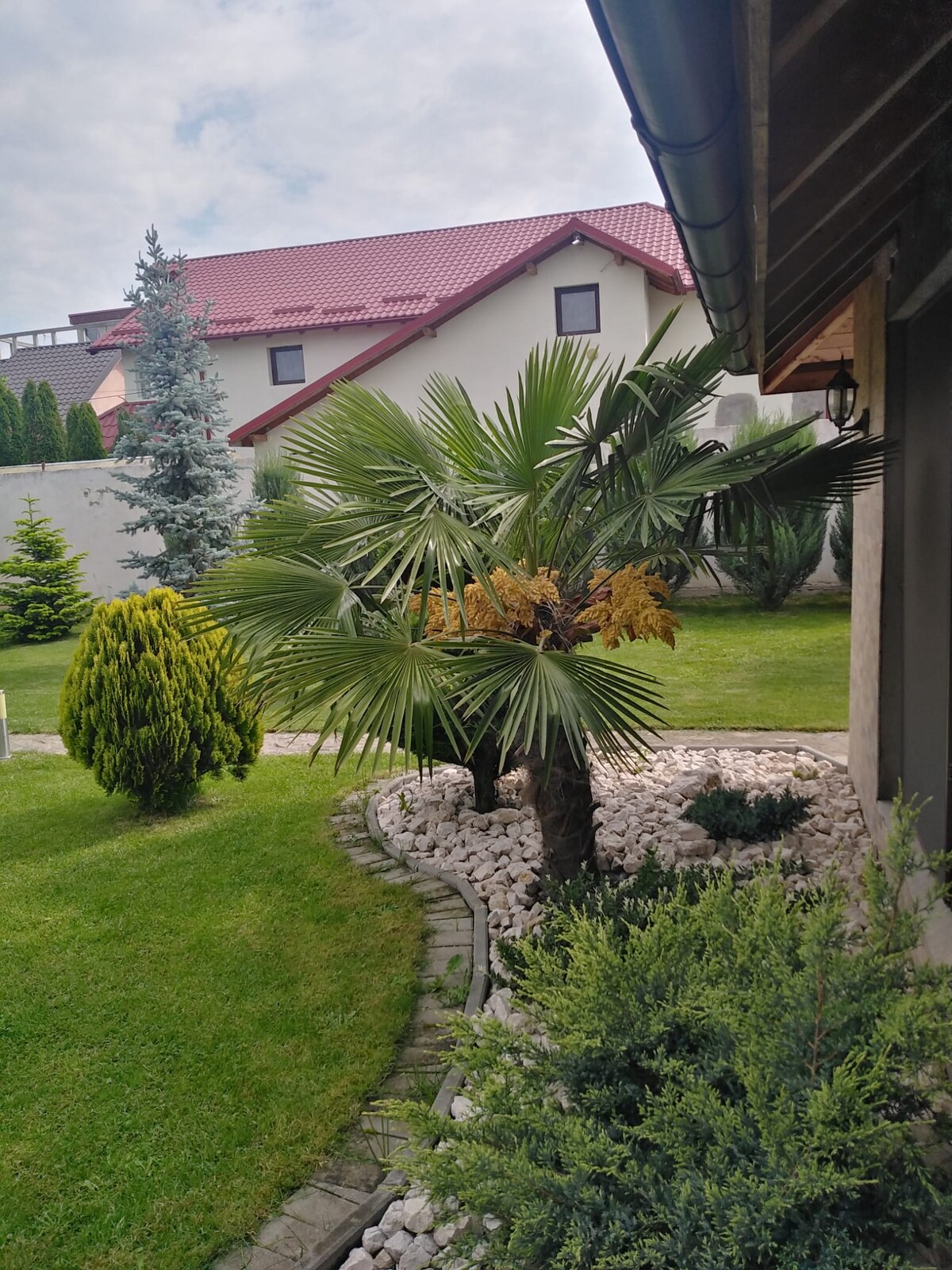 A landscaped garden features lush green lawns interspersed with a variety of plants, including a palm tree and manicured shrubs. A stone pathway winds through the area, leading towards a white gravel section. In the background, a double-storied building with a red roof is visible.