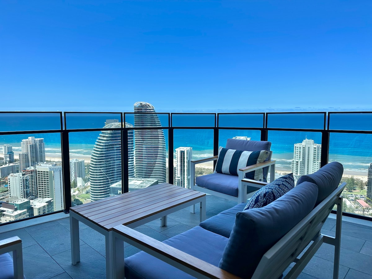 An outdoor balcony features comfortable lounge seating and a wooden coffee table, offering panoramic views of the ocean and city skyline. The clear blue sky enhances the scenic landscape, highlighting the vibrant coastline and nearby high-rise buildings.