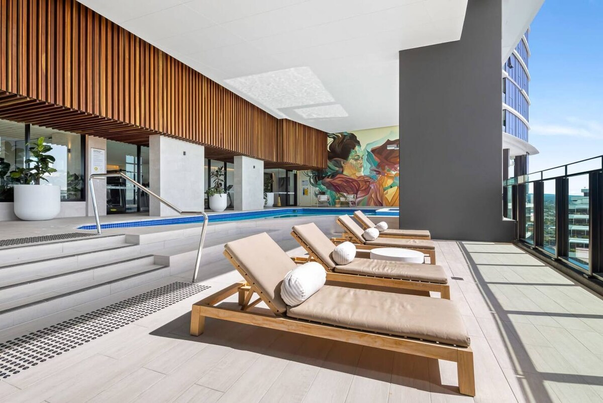 A modern outdoor lounge area is depicted, featuring several wooden sun loungers positioned beside a partially indoor heated pool. The backdrop includes a wall adorned with colorful artwork, while large windows provide ample natural light and views of the surrounding area.