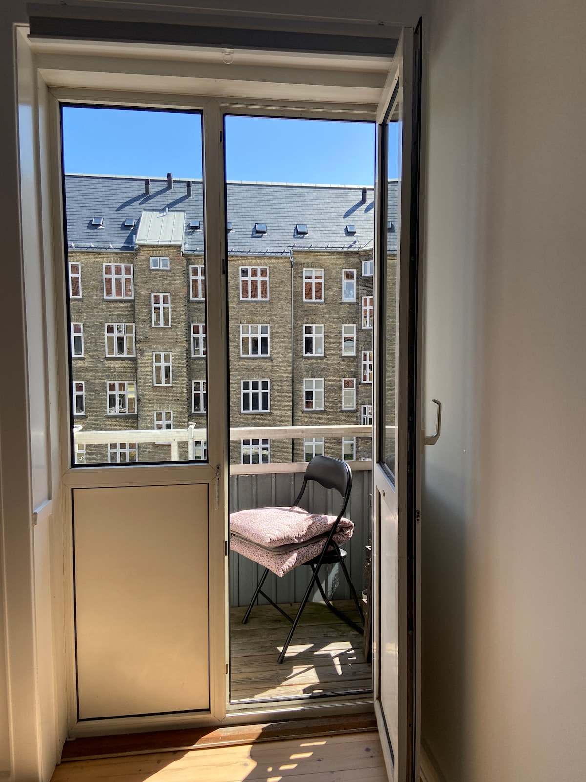 A view from an interior doorway opens to a small balcony. A single chair sits on the wooden deck, providing a place to enjoy the outside. Large windows from adjacent buildings are visible across the street, under a clear blue sky.