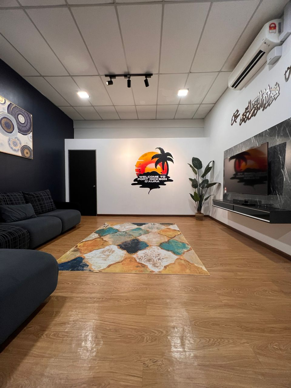 A spacious living area is presented with a large sectional sofa, arranged to face a modern entertainment unit. A decorative area rug featuring various colors lies on the polished wood floor. A mural of a sunset with palm trees adorns the wall, enhancing the room's ambiance.