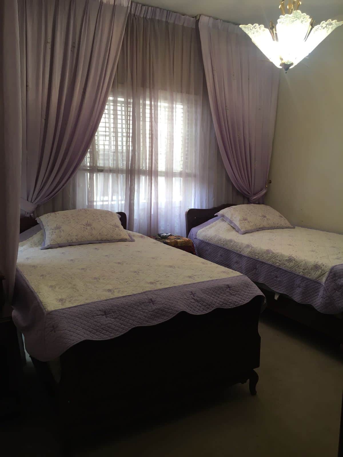 A serene guest room is presented with two twin beds, each topped with light purple bedspreads and matching decorative pillows. Soft, sheer curtains filter natural light from the window, and a delicate chandelier hangs above, creating a gentle ambiance.