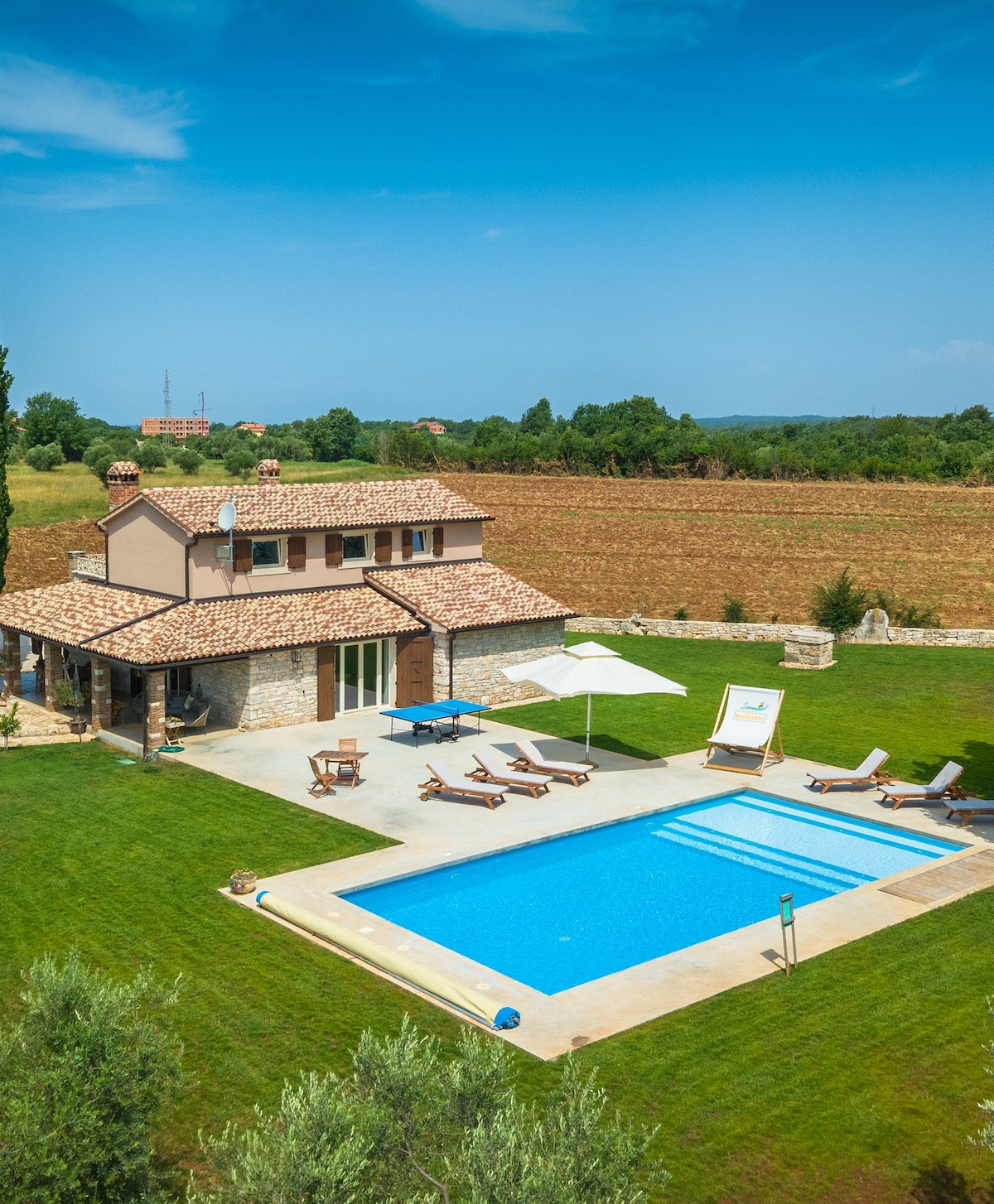A two-story villa is shown in a serene countryside setting, featuring a clear blue pool surrounded by comfortable lounge chairs. Lush greenery and a stone patio create an inviting space for relaxation. The rustic roof adds character to the charming architecture.
