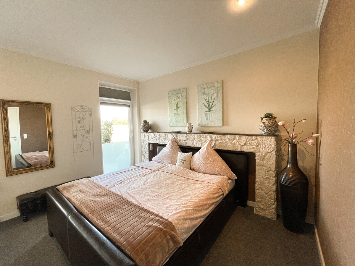 A spacious bedroom features a comfortable bed with plush pillows and a decorative throw. Elegant artwork adorns the walls, while a large mirror reflects the room's design. A window offers access to a private balcony, allowing natural light to brighten the space.