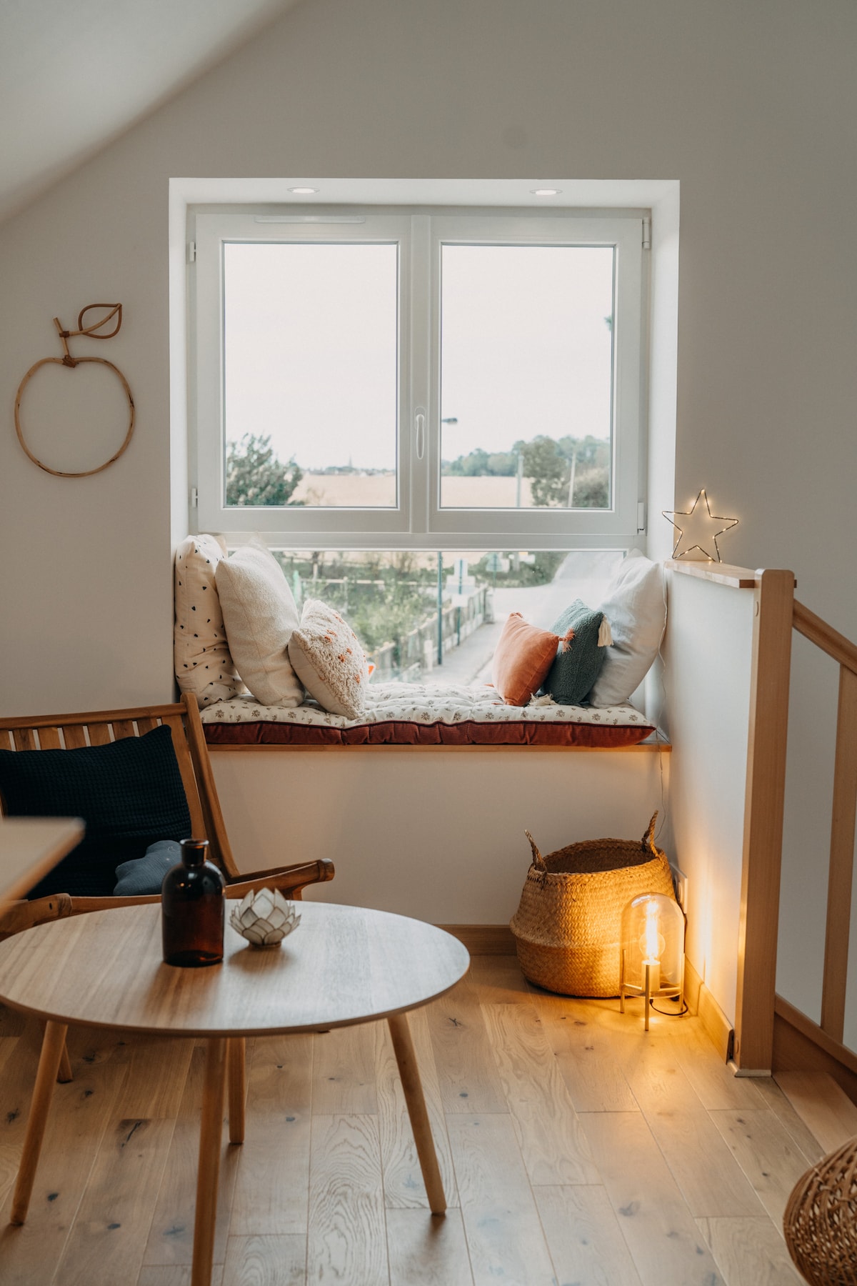 A cozy nook is featured near a large window, offering a view of the surroundings. Cushions sit comfortably on a window seat, complemented by a woven basket and a low wooden table. Soft lighting from a lamp adds to the welcoming ambiance.