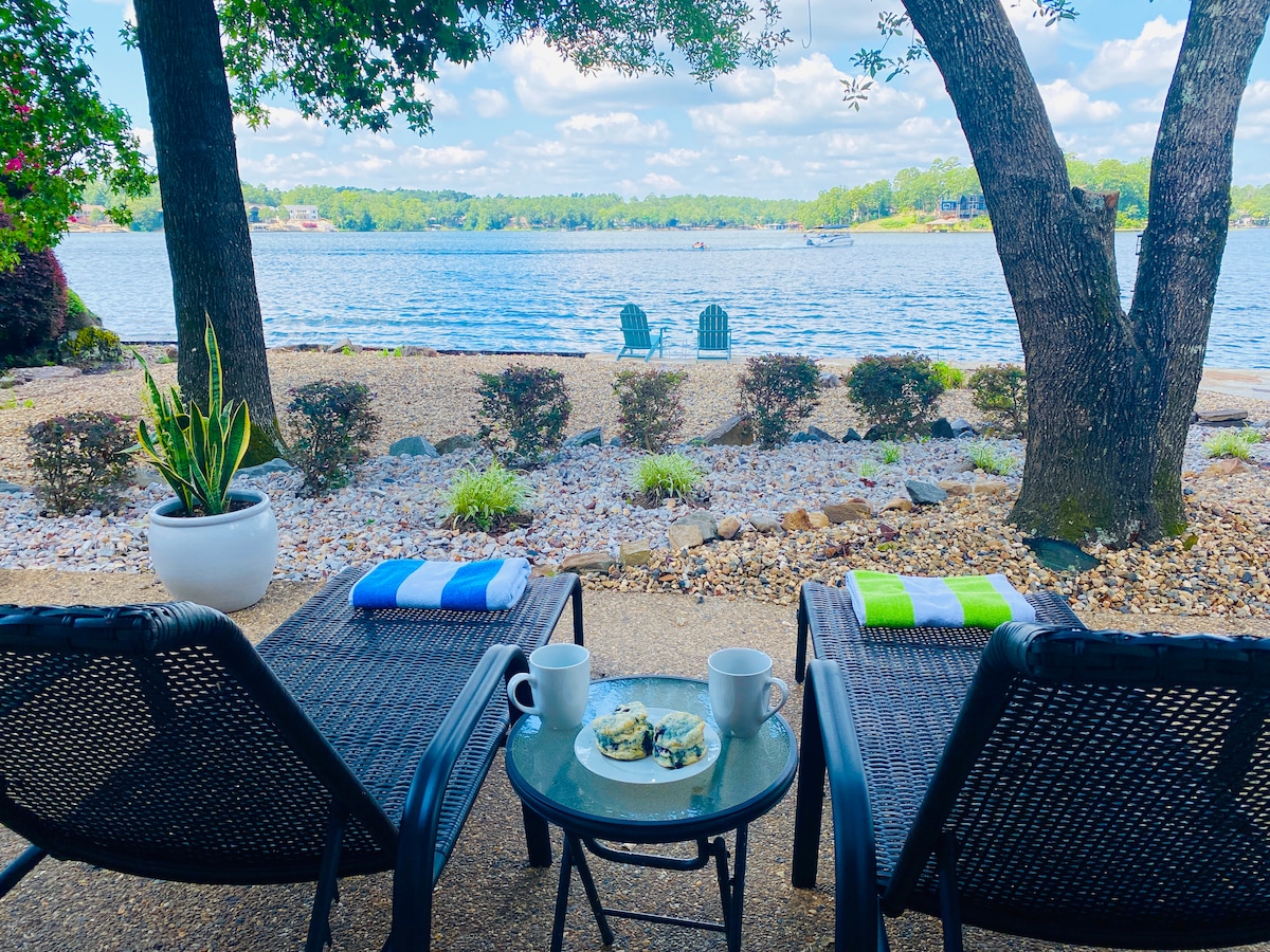 Two lounge chairs are positioned on a patio, facing a scenic view of Lake Balboa. A small table displays two cups and a plate of food. The shoreline features gentle ripples in the water, with greenery and a few seating areas visible in the distance.