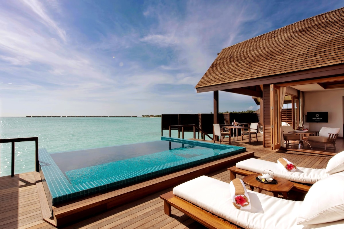 A private pool overlooks calm turquoise waters, surrounded by a wooden deck. Two sun loungers are positioned nearby, featuring plush cushions. A dining area can be seen in the background, ideal for enjoying meals with a picturesque view.