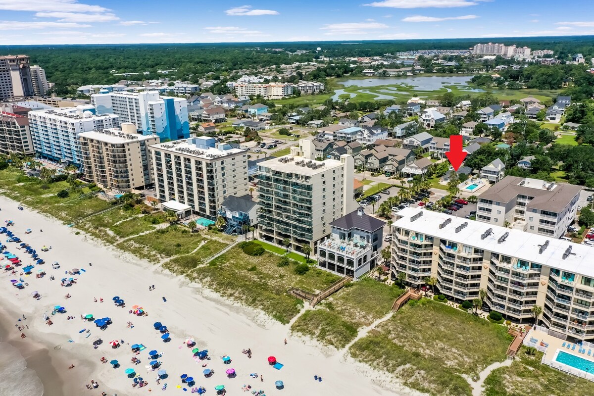 Aerial view of a beachfront area showing multiple structures along the shoreline and clusters of umbrellas on the sand. The proximity to the beach is highlighted, with a small pool visible in the background surrounded by greenery.