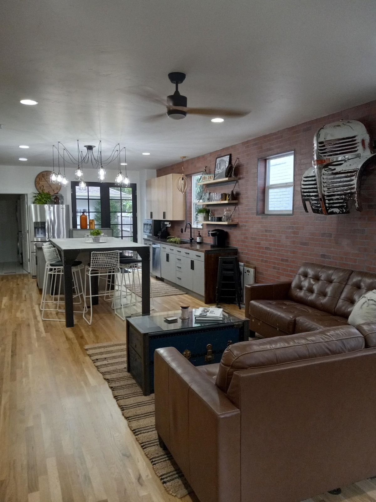 An open-concept living area features a modern design with a cozy leather sofa and an accent chair. A rustic coffee table sits on a textured rug, with an industrial-style dining area visible in the background. Large windows and ceiling lights enhance the natural lighting.