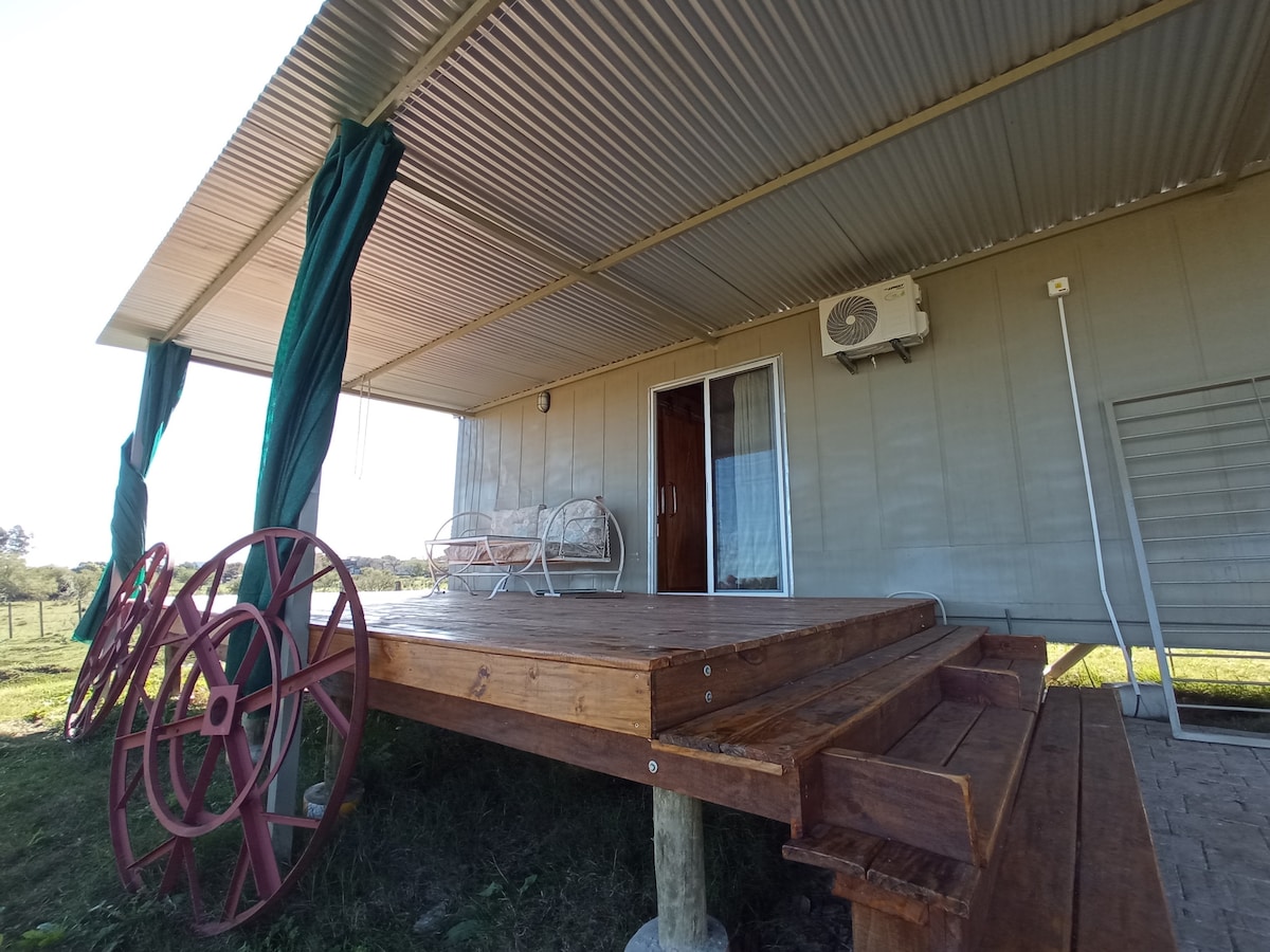 A spacious wooden deck is featured in front of a light gray house, offering a welcoming seating area with a bench. An air conditioning unit is installed on the wall, while green curtains sway gently in the breeze. Views of the surrounding landscape are visible beyond the deck.