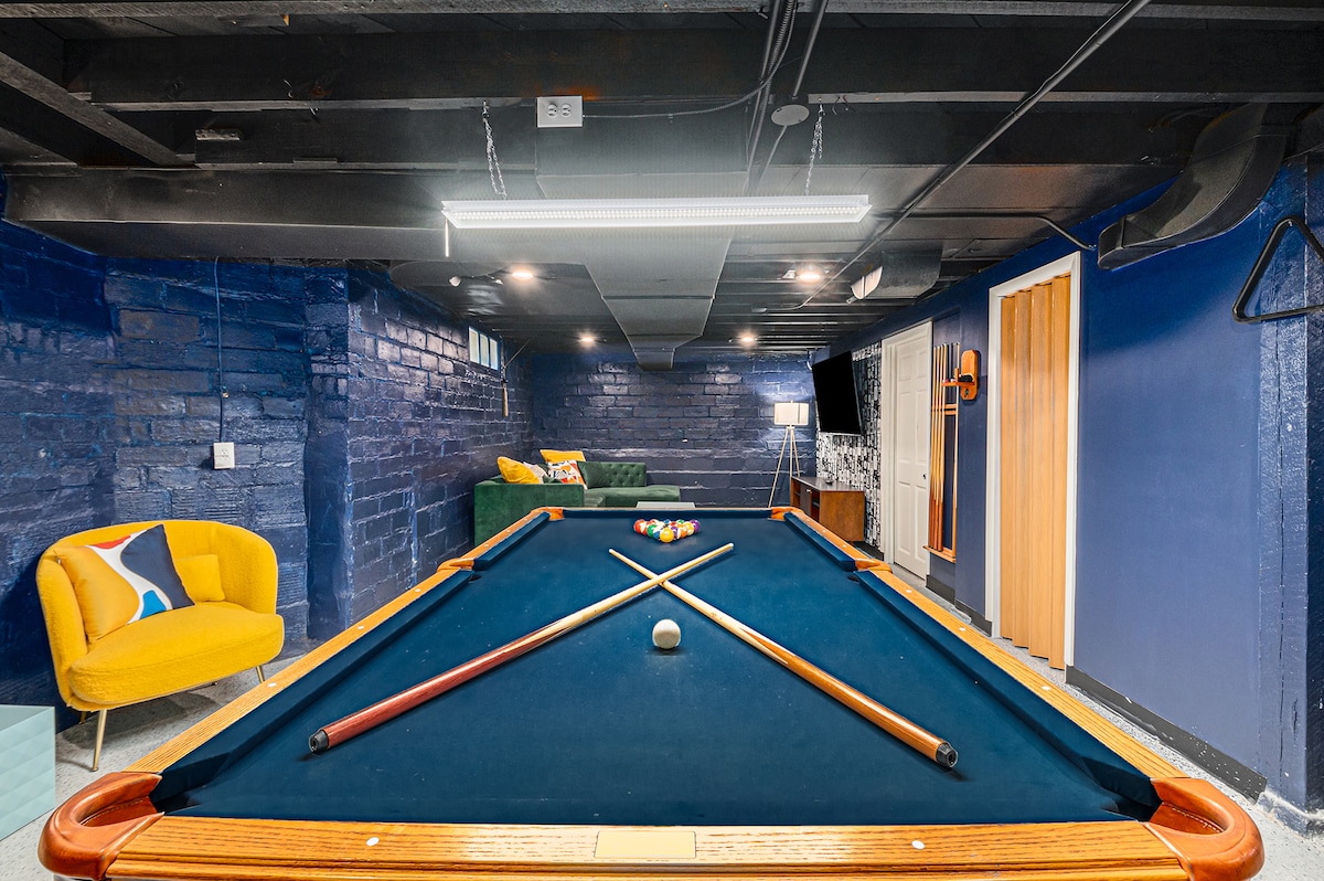 A game room is highlighted by a competition-grade pool table, featuring chalk and cues positioned atop its blue felt. Cozy seating is visible in the background, with vibrant accents against the dark walls, creating a warm and inviting gaming atmosphere.