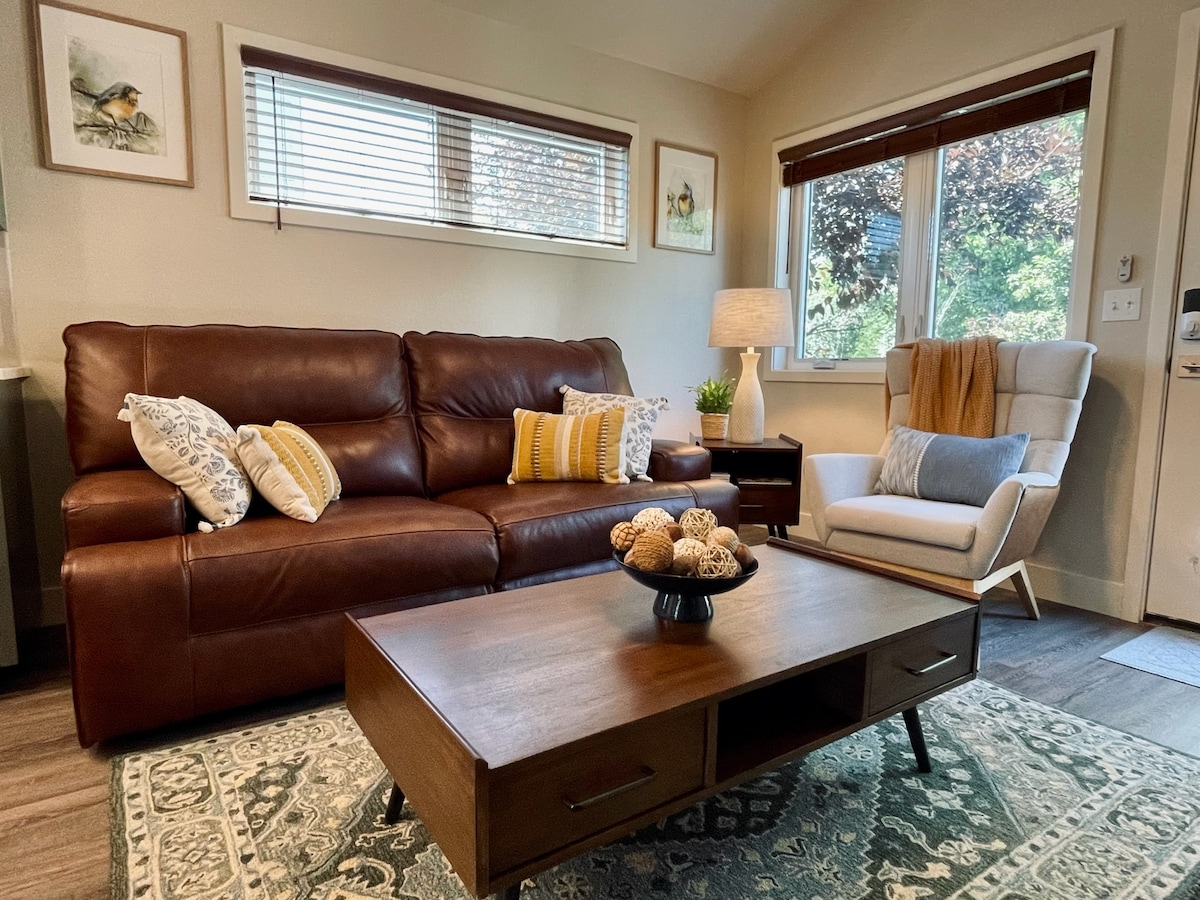 A cozy living area is presented with a leather sofa adorned with throw pillows, positioned beside a bright window. A light-colored armchair complements the space, along with a wooden coffee table featuring a decorative bowl. Warm tones are balanced by a patterned area rug.