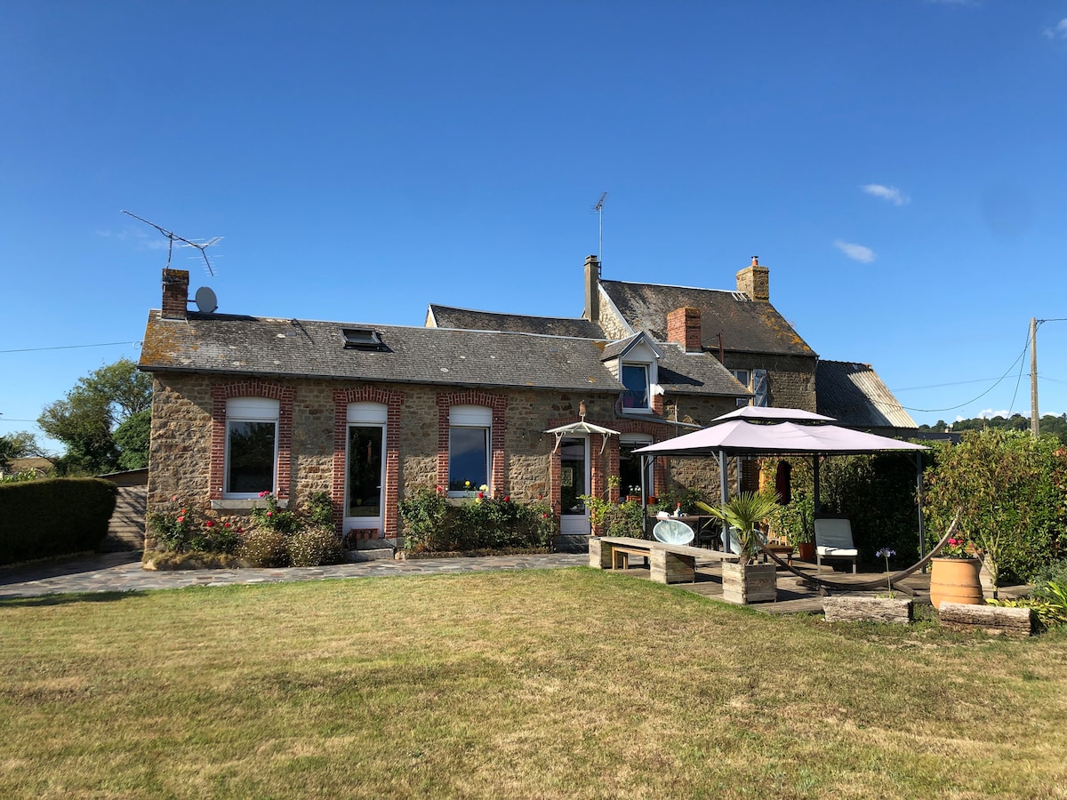 A countryside cottage is depicted, featuring a mix of stone and brick exteriors. A spacious lawn surrounds the structure, complemented by a shaded seating area under a canopy. Nearby, flowering plants add color to the landscape, with clear blue skies above.