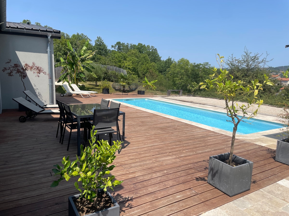 An outdoor terrace features a wooden deck surrounding a swimming pool. Several potted plants are placed around the area. Dining furniture, including a table and chairs, is visible, alongside two lounge chairs positioned beside the pool, all under a clear blue sky.