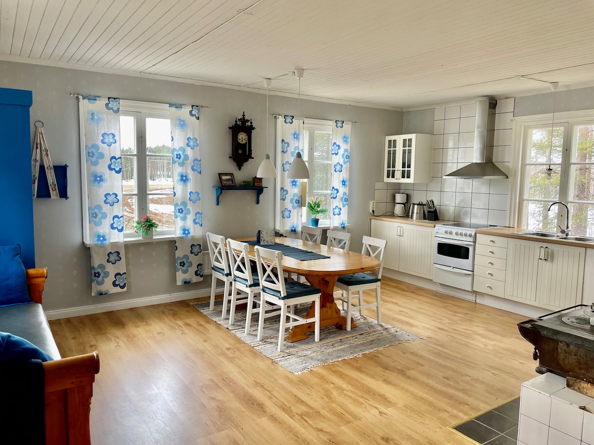 A bright kitchen and dining area is visible, featuring a wooden dining table surrounded by six white chairs. Windows adorned with blue floral curtains allow natural light to fill the space, complemented by light wood flooring and white cabinetry.