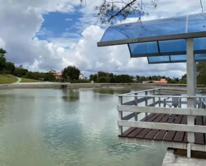 A covered wooden deck extends over calm water, providing a serene view of the surrounding landscape. Clear skies are visible above, with gentle clouds adding depth to the scene, while lush greenery lines the far bank.