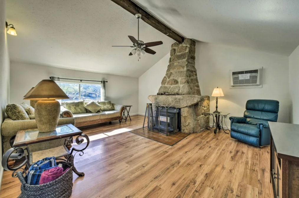 A welcoming living area features a cozy stone fireplace and comfortable seating, illuminated by natural light through the large window. A ceiling fan adds a cooling element, while a side table holds a lamp, providing additional light. Wood flooring enhances the warm ambiance.