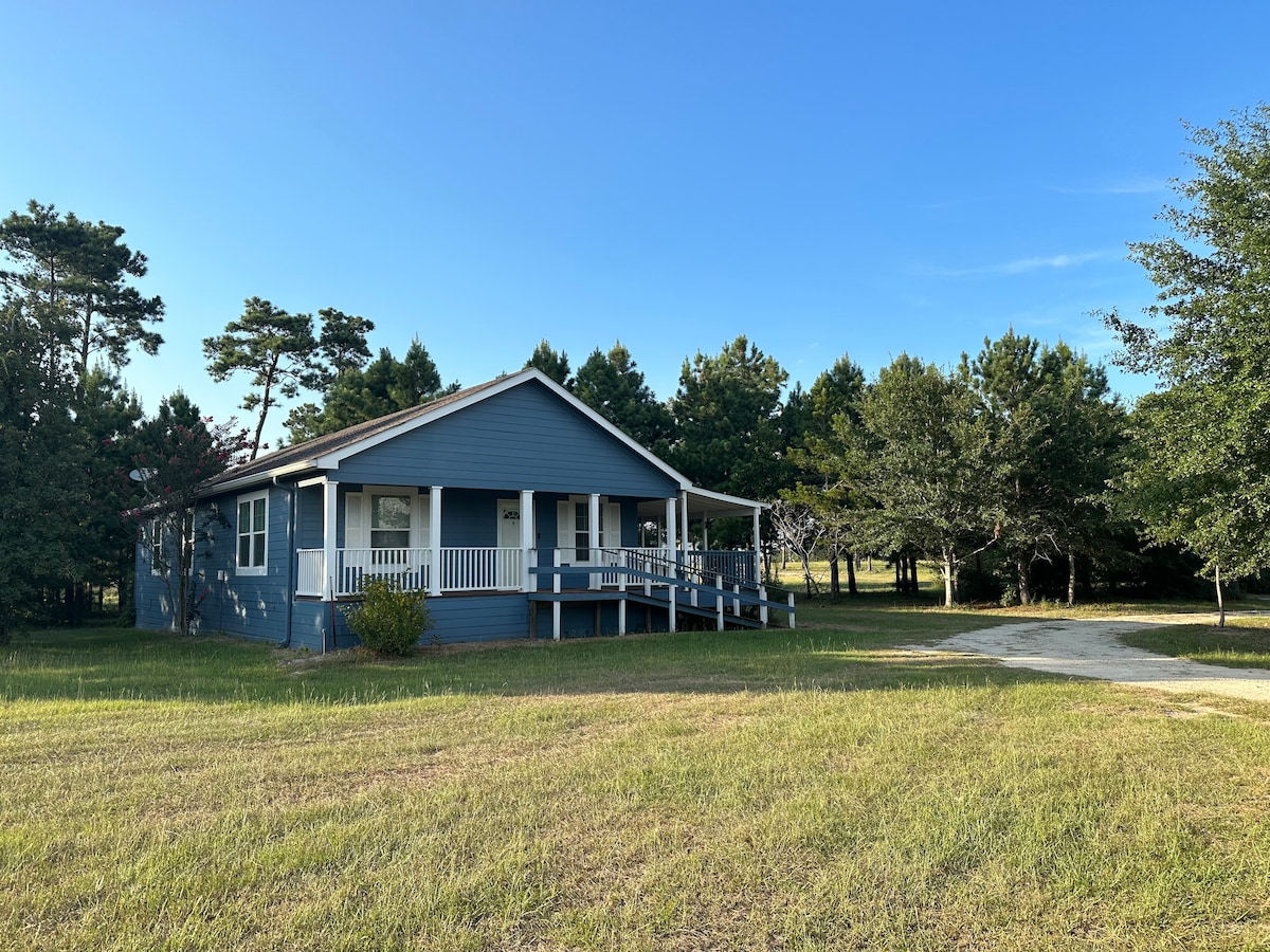 A single-story blue house is set on a grassy lot surrounded by trees. A covered front porch with white railings offers a welcoming entrance. The well-maintained yard is spacious, providing a serene outdoor environment.